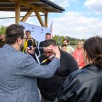 Mężczyzna zakłada medal innej osobie na zewnątrz. W tle kilka osób na zielonym tle oraz niebieskie niebo z chmurami. Scena odbywa się przed drewnianą wiatą.