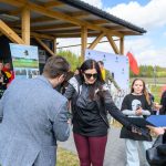 Grupa ludzi na zewnątrz, kobieta otrzymuje nagrodę. Drewniana konstrukcja w tle, widoczna polska flaga. Słoneczny dzień, niebo z białymi chmurami.