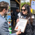 Na zdjęciu kobieta w okularach przeciwsłonecznych trzyma dyplom podziękowania. Mężczyzna wręcza jej nagrodę. Tło jest rozmyte, z ludźmi oraz banerami.