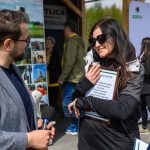 Na zdjęciu dwoje ludzi rozmawia na zewnątrz. Kobieta w czarnej kurtce trzyma tablicę z tekstem. Tło jest zielone, z plakatami i ludźmi.