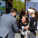 Mężczyzna w marynarce podaje rękę dziewczynce na tle plakatów. Obok stoi osoba z opaską na oczach, przy drewnianym płocie. Dzień jest słoneczny, z zielenią w tle.