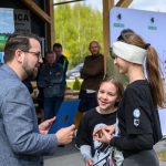 Na zdjęciu mężczyzna w okularach wręcza dokument dziewczynce w czerni. Tło to przyroda i namiot promocyjny z logo. Kolory dominujące to niebieski, zielony i czarny.