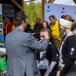 Na zdjęciu mężczyzna zakłada medal na szyję dziewczynce. Tło zdobią banery z plakatami w zielonych kolorach. Widać też grupę ludzi w tle.