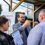 Na zdjęciu trzy osoby uczestniczą w ceremonii wręczenia medalu. Mężczyzna wręcza medal, kobieta trzyma dyplom. W tle widać plakat i zielone drzewa.