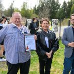 Nie wiem, kto jest na zdjęciu. Trzy osoby na zielonym trawniku, jedna trzyma podziękowanie. Tło to drzewa i ogrodzenie.