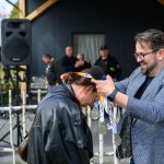 Mężczyzna wręcza kobiecie medale, oboje uśmiechnięci. W tle plakat Mazowsze, ludzie i sprzęt nagłaśniający. Dominują kolory czarny i niebieski.