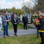 Grupa ludzi stoi na zewnątrz na chodniku. Kobieta wręcza bukiet kwiatów. W tle widać drzewa i zaparkowane auta.