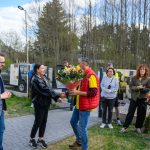 Grupa ludzi na zewnątrz, mężczyzna wręcza kobiecie bukiet kwiatów. Tło to drzewa i ogrodzenie. Ubrania w żywych kolorach.
