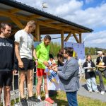 Na zdjęciu trzech mężczyzn na podium otrzymujących nagrody. Ubrani w sportową odzież, w tle drewniana altana i niebo z chmurami. Obok stoją dzieci z pucharami.