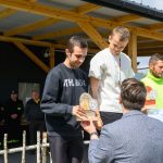Trzej mężczyźni z trofeami na podium. Jeden jest w czarnej bluzie, drugi w białej koszulce, trzeci w zielonej kurtce. W tle drewniane zadaszenie i osoby w ubraniach z napisem STRAŻ.