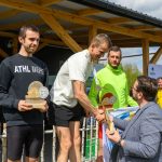 Trzech mężczyzn na podium, trzymają trofea. Jeden w jasnej koszulce ściska rękę organizatora. Tło z drewnianym zadaszeniem i flagą Polski.