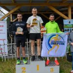 Na podium stoją trzej mężczyźni z nagrodami. Tło stanowi drewniana konstrukcja, obok banery i zieleń. Dwaj trzymają flagę z napisem MIĘSKI KLUB BIEGACZA DREPTAK.