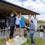 Na podium stoją trzy kobiety, z których jedna otrzymuje nagrodę. Kobiety ubrane są w sportowe stroje, dominują kolory czarny i niebieski. W tle widać zadaszoną scenę i grupę obserwujących osób.