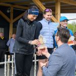 Trzy kobiety na podium otrzymują nagrody od mężczyzny. Ubrania są w ciemnych odcieniach, tło to drewniana konstrukcja. Obserwatorzy stoją w tle.