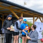 Na podium trzy kobiety otrzymują trofea. Mają sportowe stroje, jedna w niebieskiej czapce. W tle drewniana konstrukcja i baner.