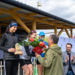 Na podium stoją trzy kobiety, jedna trzyma puchar, druga kwiaty. Tło to drewniana konstrukcja i błękitne niebo. Dookoła są klaskający widzowie.