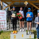 Na podium stoją trzy kobiety z trofeami, uśmiechają się. Tło tworzy drewniana altanka i baner „Mazowsze”. Wiosenna sceneria z kwiatami.