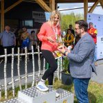 Kobieta w czerwonej kurtce na podium, w tle osoby i afisz. Mężczyzna wręcza jej nagrodę na świeżym powietrzu.