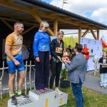 Na podium stoją trzy osoby w sportowych strojach podczas ceremonii wręczenia nagród. W tle drewniana wiata i biały baner na łące. Mężczyzna wręcza kobiecie trofeum, obok stoją dwie młode osoby z medalami.