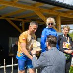 Na zdjęciu mężczyzna w sportowym stroju odbiera trofeum. W tle drewniana konstrukcja, słońce i zielone drzewa. Obok stoją dwie kobiety oraz kilka osób w tle.