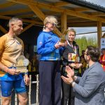 Na zdjęciu są osoby stojące na podium i odbierające nagrody. Tło to drewniana altana i zielone drzewa. Osoby mają medale i statuetki.