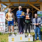 Trzy osoby na podium, trzymają puchary. Tło z drewnianą konstrukcją i plakatem Mazowsze. Słoneczny dzień, trawa i niebo.