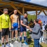 Trzech mężczyzn na podium, w sportowych strojach, z medalami. Tło to drewniana konstrukcja i roślinność. Osoba wręczająca nagrody, w okularach i marynarce.