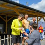 Zawodnicy na podium otrzymują nagrody na zewnątrz. Mężczyzna w żółtej bluzie przyjmuje trofeum. W tle konstrukcja z drewna i niebo z chmurami.