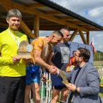 Na zdjęciu czterech mężczyzn; jeden wręcza trofea. W tle drewniana wiata i niebo. Przewaga jasnych kolorów.