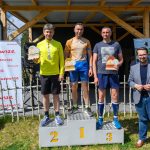 Trzech mężczyzn na podium z trofeami; jeden w żółtej koszulce, drugi w beżowo-niebieskiej, trzeci w granatowej. Tło z banerami na zewnątrz, jasnozielona trawa. Stoją obok nich mężczyźni w maryn