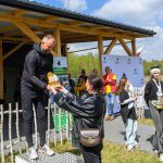 Na zdjęciu mężczyzna wręcza nagrodę kobiecie na zewnątrz drewnianego budynku. Tło jest zielone z drewnianym płotkiem, kilka osób w tle. Atmosfera jest słoneczna i radosna.