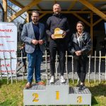Troje ludzi stoi na podium: miejsca 1, 2, 3. Ubrani w ciemne kurtki, otoczeni drewnianym ogrodzeniem, z banerem Mazowsze w tle.