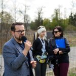 Mężczyzna w okularach przemawia do mikrofonu na tle siatki. Obok dwie kobiety, jedna z teczką. Zielone drzewa w tle.