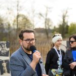 Mężczyzna z brodą mówi przez mikrofon, ubrany w szarą marynarkę. W tle dwie kobiety i plakaty. Jasne, naturalne otoczenie z drzewami.