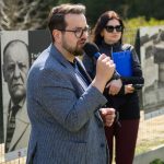 Mężczyzna w okularach trzyma mikrofon na tle ogrodzenia z czarno-białymi zdjęciami. Kobieta w okularach stoi za nim. W tle zielone drzewa.