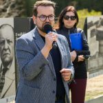 Nie rozpoznaję osób na zdjęciu. Mężczyzna w kraciastej marynarce mówi do mikrofonu. W tle kobieta z niebieską teczką i zielone drzewa.