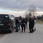 Na zdjęciu grupa osób stoi obok czarnego vana na wiejskiej drodze. W tle widać drzewa i altankę pod pochmurnym niebem. Ludzie są zajęci rozmową i notowaniem.