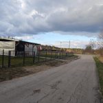 Droga wiejska prowadząca obok budynku z dachem i polską flagą. Po lewej trawa i metalowe ogrodzenie. Niebieskie niebo z chmurami.