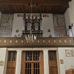 POPmPzu_ Kościelny chór z organami oraz drewnianym balkonem, zdobiony złotymi wzorami. Tło w neutralnych kolorach z religijnymi napisami. Po bokach widać dekoracje ścienne i flagi.