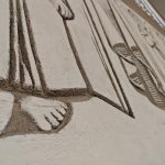 TfRKzfEc Płaskorzeźba na ścianie przedstawia fragment ludzkiej postaci. Dominują beżowe i brązowe odcienie. Ujęcie z bliska ukazuje fakturę i szczegóły reliefu.