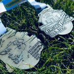 Medale leżą na zielonej trawie. Są srebrzyste, z wyrytymi napisami i wzorami. Widoczna jest również biała wstążka.