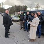 Grupa osób stoi przy czarnym aucie na wiejskiej drodze. Kobieta w jasnym płaszczu trzyma dokumenty. Tło: kilka aut, drzewa i pochmurne niebo.