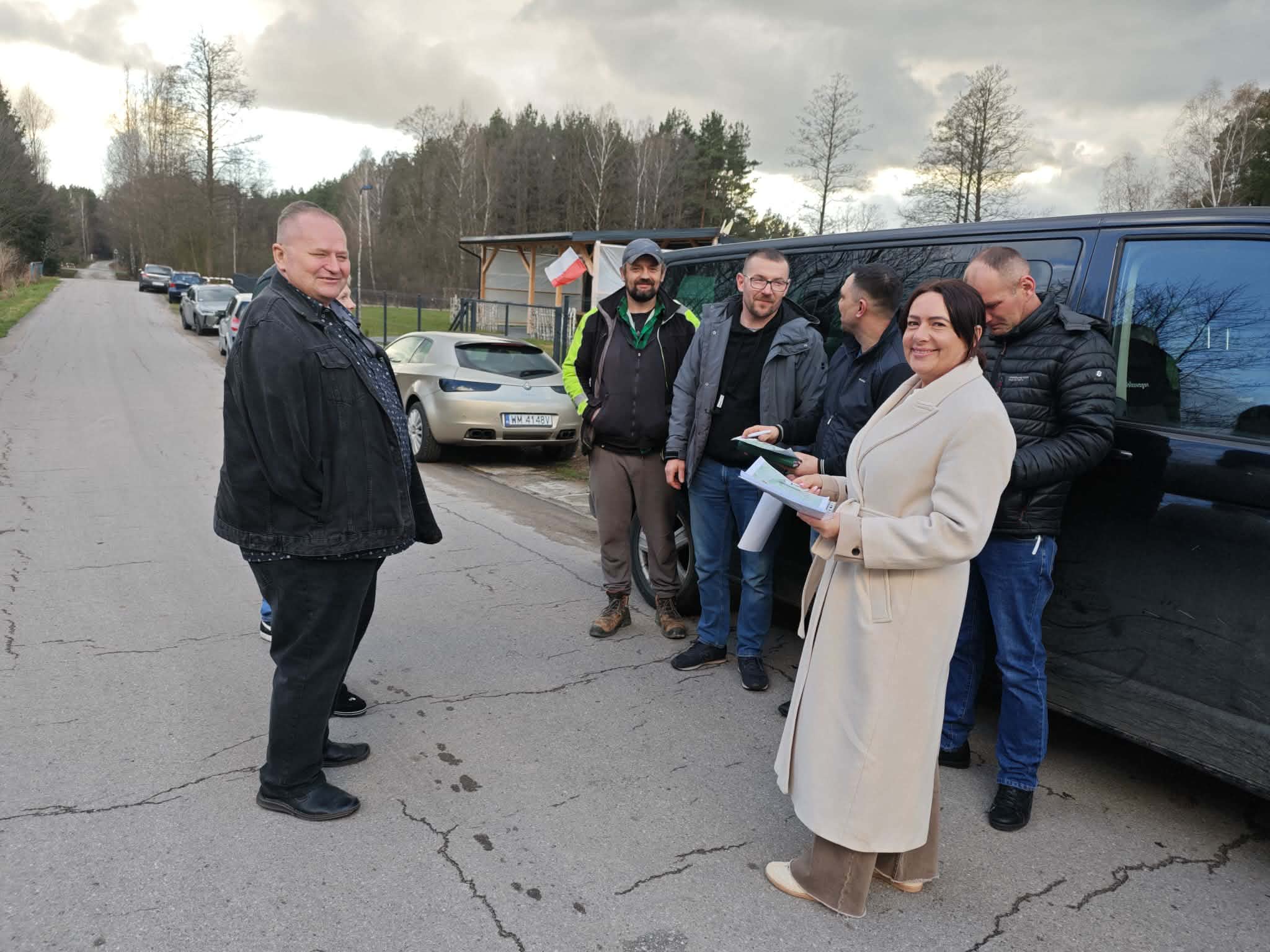 Grupa osób stoi przy czarnym aucie na wiejskiej drodze. Kobieta w jasnym płaszczu trzyma dokumenty. Tło: kilka aut, drzewa i pochmurne niebo.