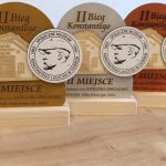 Nagrody z drewna na II Bieg Konstantego w różnych kolorach: brąz, szary, pomarańczowy. Widać nazwę muzeum i datę. Tło jasne, stonowane.