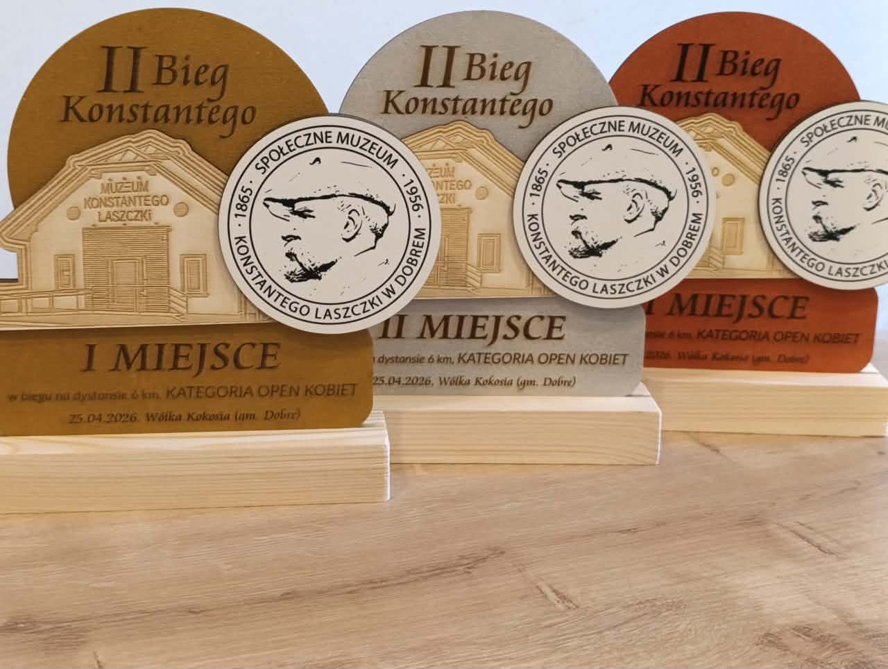 Nagrody z drewna na II Bieg Konstantego w różnych kolorach: brąz, szary, pomarańczowy. Widać nazwę muzeum i datę. Tło jasne, stonowane.