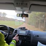 Samochód na polnej drodze w lesie. Kierowca w żółtej kurtce trzyma kierownicę. Na desce rozdzielczej mapa i ekran nawigacji.