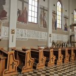 bPaigTjN Drewniane ławki z krzyżami w kościele; na ścianach freski i witraże. Jasne wnętrze z kafelkami na podłodze.