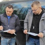 Dwóch mężczyzn stoi przy czarnym samochodzie, analizując dokumenty. Noszą ciemne kurtki i dżinsy. W tle widać odbicie chmur i drzew.