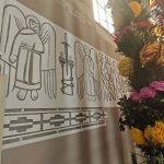 cVQ9oLQ8 Malowidło przedstawia anioły w brązowych konturach na jasnej ścianie. Obok jest kolorowa palma z kwiatami w odcieniach żółtym i różowym. Tło ma beżowe odcienie z fragmentem tekstu.