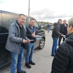 Na zdjęciu kilku mężczyzn stoi przy ciemnym vanie, z teczkami w ręku, na tle chmur i drzew. Noszą ciemne, casualowe kurtki. Tło urbanistyczne, w oddali widać samochody.